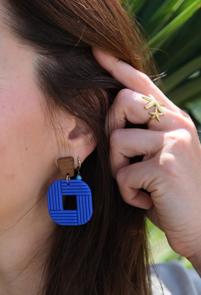 Découvrez Tara, des boucles d’oreilles pendantes qui associent une pièce rectangulaire en bois à un carré aux bord arrondi façonné à la main, en argile polymère bleu roi. Ultra-légères et confortables, ces boucles faites main sont conçues pour révéler votre singularité et donner du caractère à votre tenue.