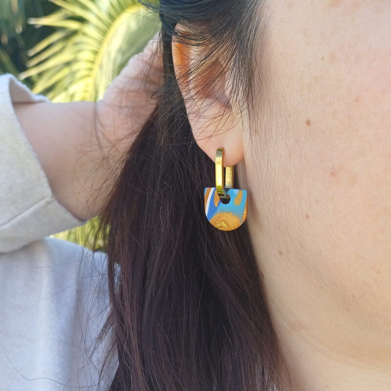 Apportez une touche de modernité à votre tenue grâce aux créoles avec pampille interchangeable, Inès. Ces boucles d’oreilles délicates se distinguent par un demi cercle marbré de différentes teintes de bleu, de blanc et de doré. Il est posé sur une créole dorée ou argentée, à choisir selon vos préférences.